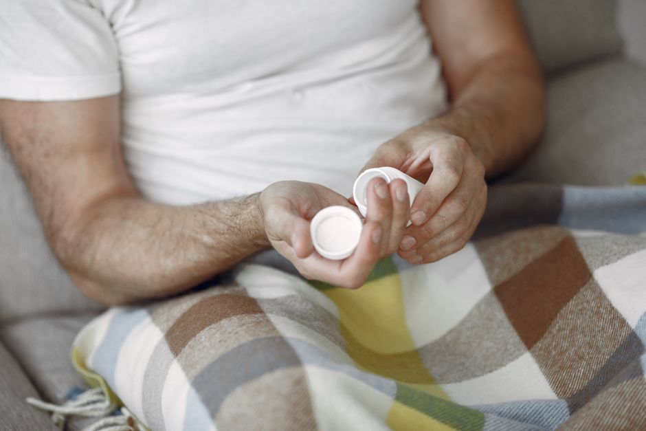 Hombre adulto sentado en el sofá, sosteniendo pastillas, envuelto en una manta acogedora, ambiente relajado.