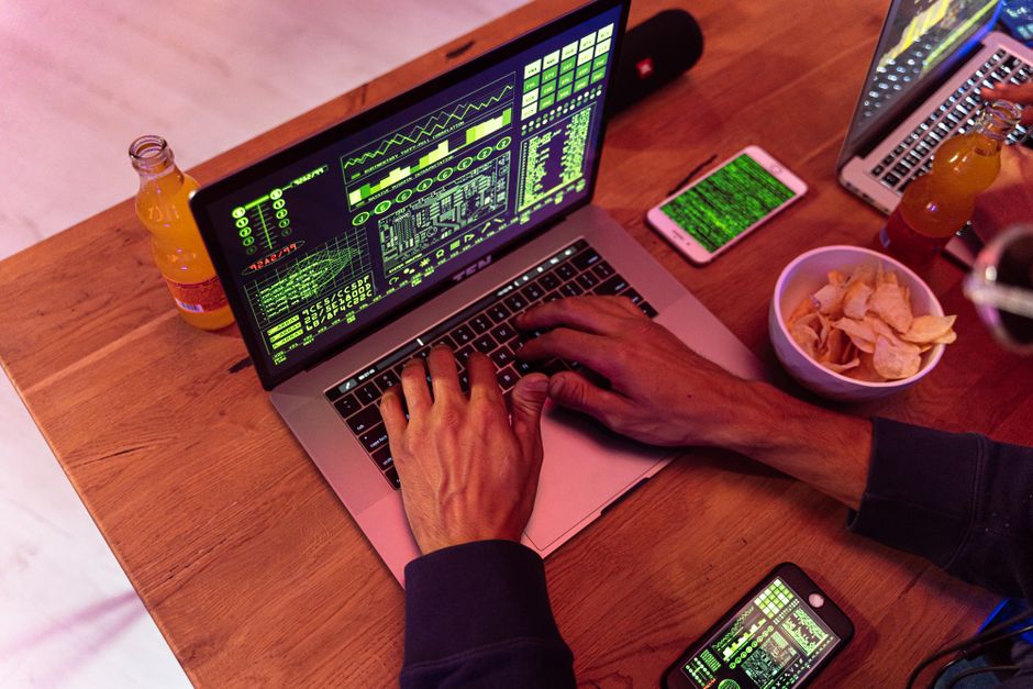 Manos escribiendo en un portátil con código, teléfono en el escritorio, simbolizando ciberseguridad.