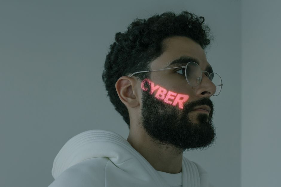Hombre con barba y proyección de texto cibernético, usando gafas y sudadera blanca.