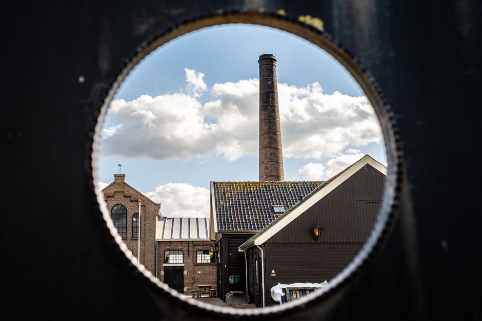 Edificio industrial histórico y chimenea vistos a través de una abertura circular bajo un cielo nublado.
