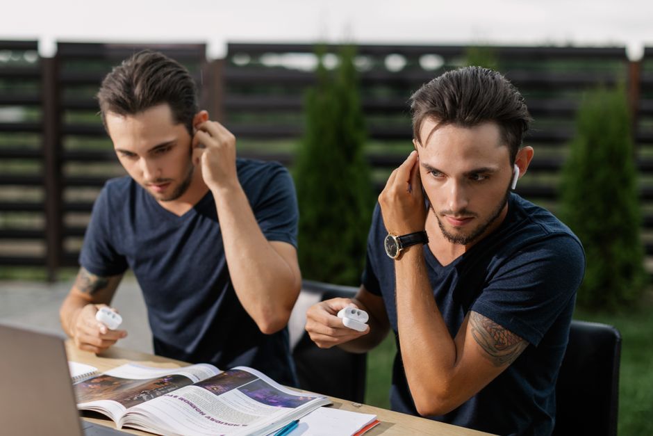 Hermanos gemelos estudiando al aire libre, usando auriculares inalámbricos con una laptop.