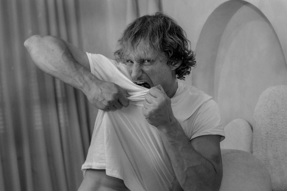 Retrato en blanco y negro de un hombre mordiendo su camisa en una pose expresiva en interiores.
