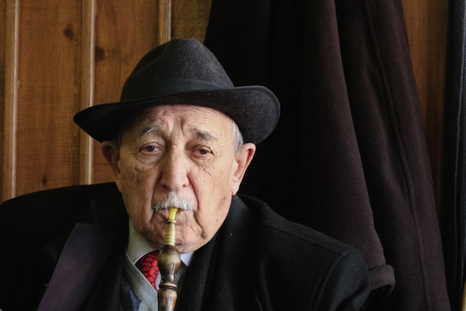 Hombre mayor con traje y sombrero fumando shisha en interiores, simbolizando ocio y jubilación.