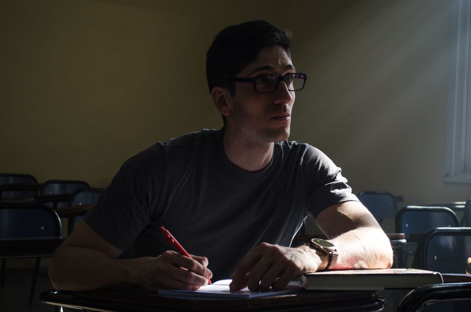 Un estudiante pensativo toma apuntes en un aula iluminada por el sol, sumido en una profunda concentración.