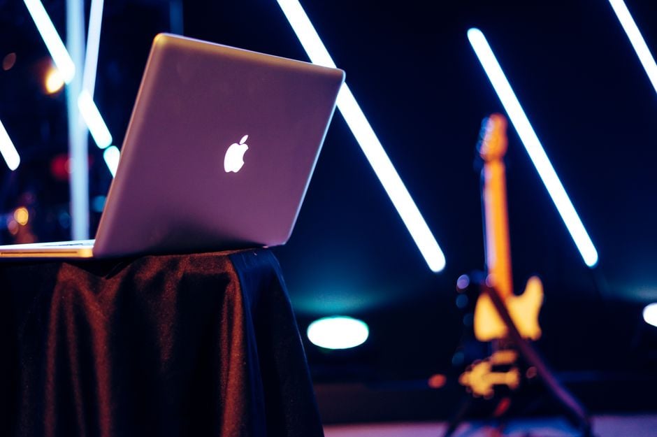 Un portátil elegante se encuentra sobre una mesa con luces de neón vibrantes y una guitarra al fondo.