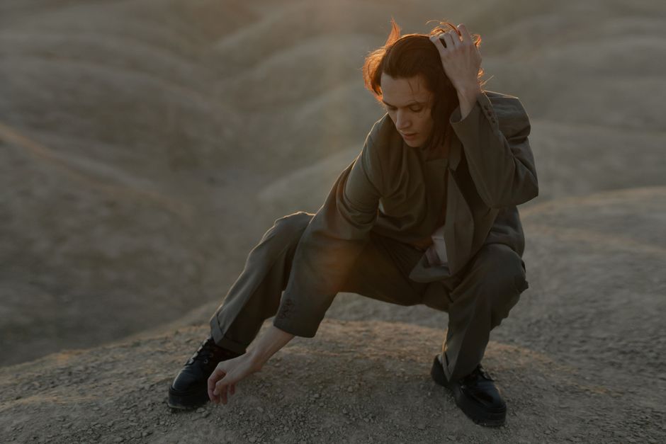 Un hombre con traje posando artísticamente en un paisaje seco y polvoriento con luz cálida de la tarde.