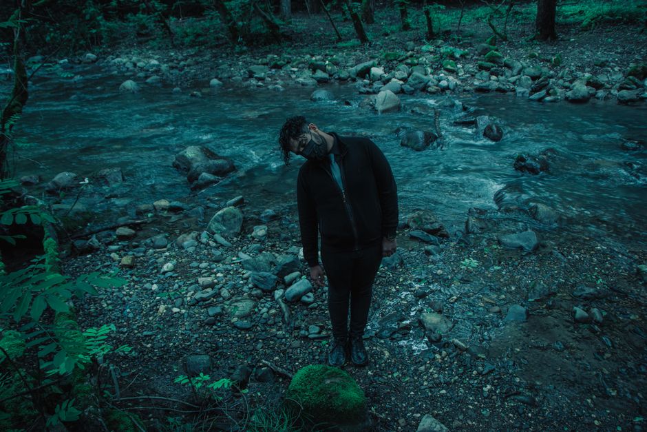 Una persona con mascarilla de pie en la orilla del río en un entorno forestal extraño.