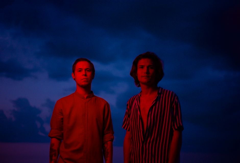 Dos hombres están bajo un cielo nocturno melancólico con nubes oscuras, iluminados por una luz azul fría y una luz roja cálida.