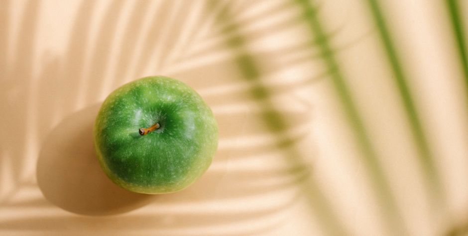 Primer plano de una manzana verde fresca con sombras de hojas de palmera bajo la luz solar, perfecta para conceptos de salud y alimentos.