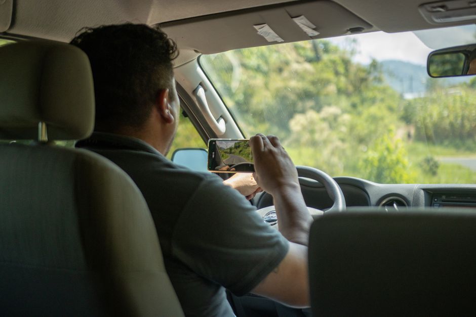 Un hombre utiliza un smartphone para capturar una escena exterior mientras conduce en el interior de un coche durante el día.