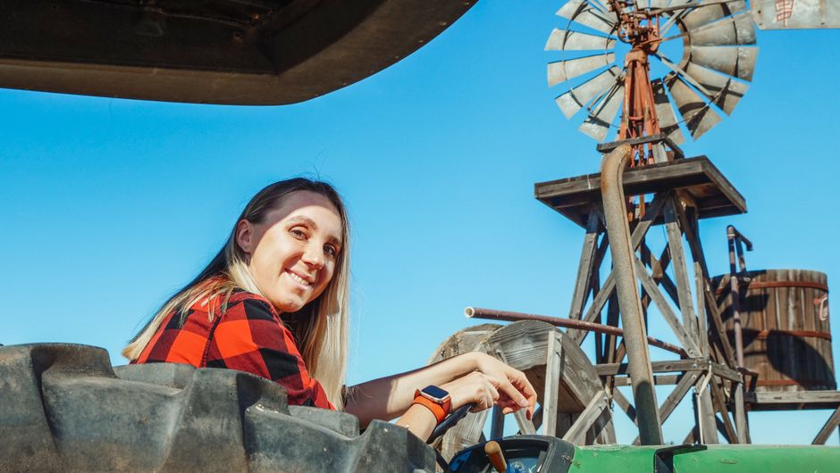 Una mujer con una camisa de cuadros sonríe desde un tractor con un molino de viento en el fondo soleado.