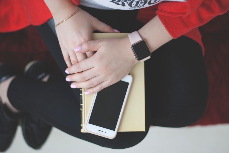 Manos de mujer con un reloj descansando sobre un libro y un smartphone, vista desde arriba.