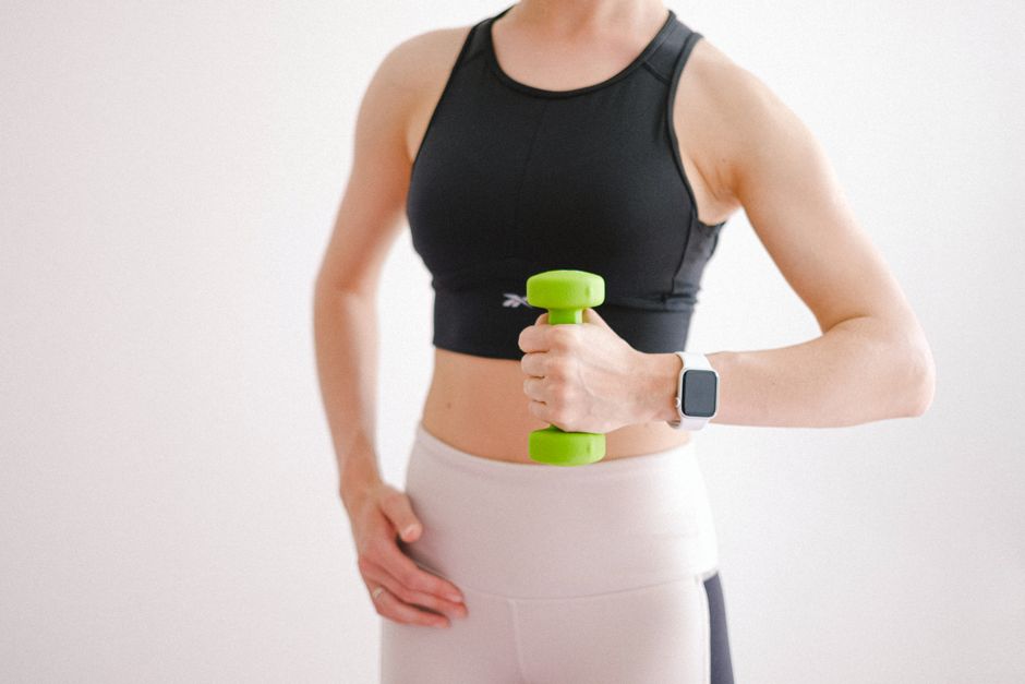 Una mujer en ropa deportiva usando una mancuerna y un smartwatch durante un entrenamiento en casa.