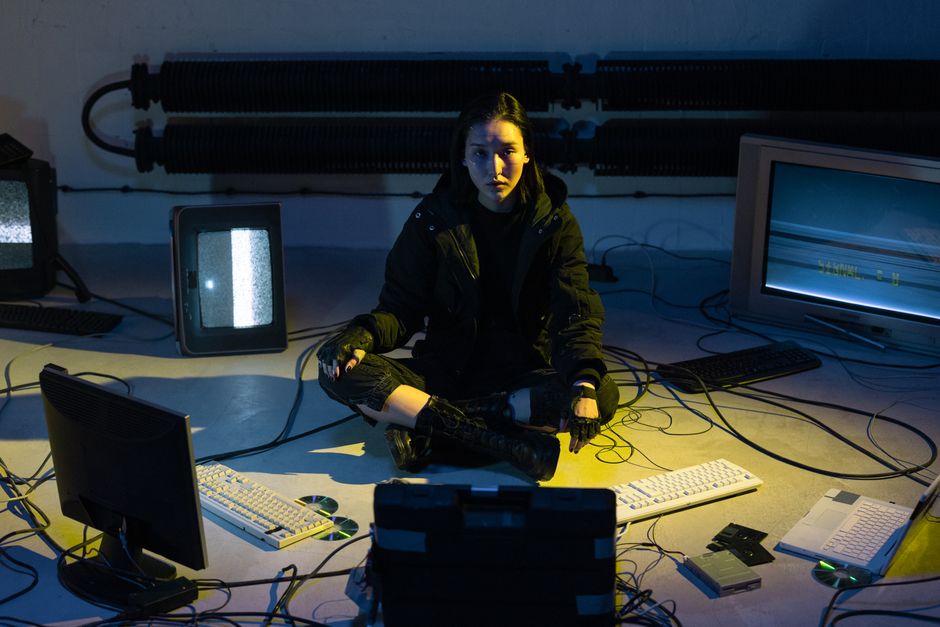 Una mujer está sentada en el suelo en una habitación tenue llena de monitores y teclados de computadora vintage.