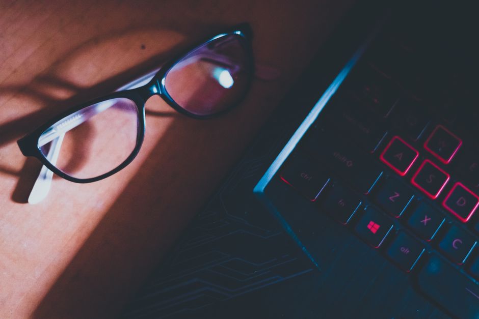 Primer plano de gafas sobre un escritorio iluminado por el sol junto a un teclado de laptop, destacando la tecnología moderna.