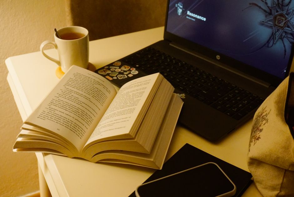 Libro abierto junto a un laptop y una taza de té en un escritorio, perfecto para estudiar o trabajar desde casa.