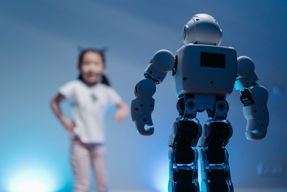 Una niña pequeña interactúa de manera juguetona con un robot humanoide en un entorno interior futurista con suave iluminación azul.