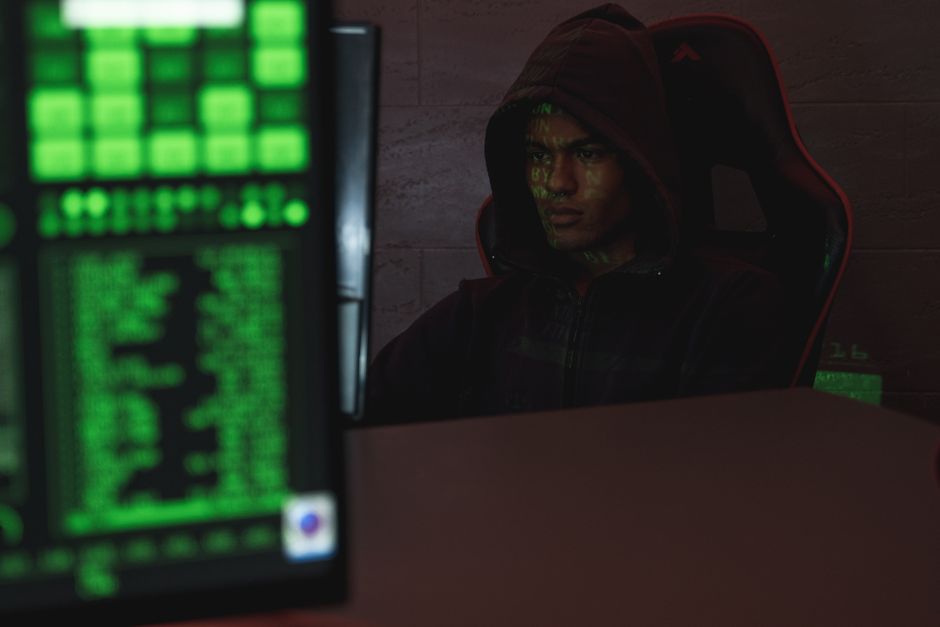 Programador con capucha enfocado intensamente en la pantalla de la computadora, asegurando la protección de datos y la ciberseguridad.