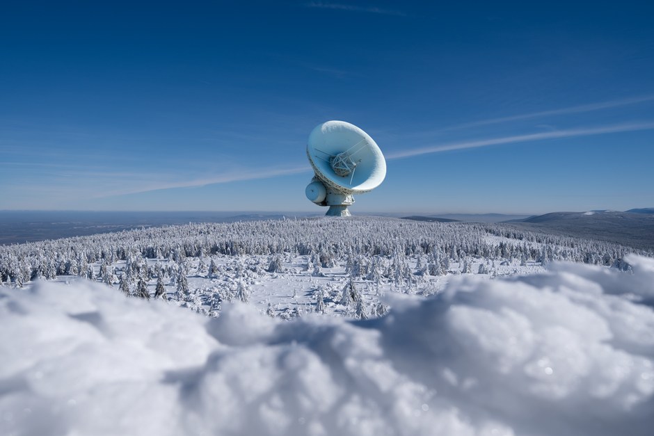 Un gran observatorio de plato satelital rodeado de un paisaje nevado y un bosque invernal bajo un cielo azul claro.