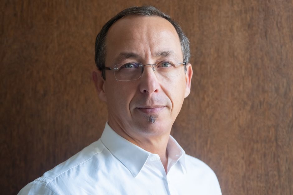 Retrato en primer plano de un hombre de mediana edad con gafas, vestido con una camisa blanca, contra un fondo de madera.