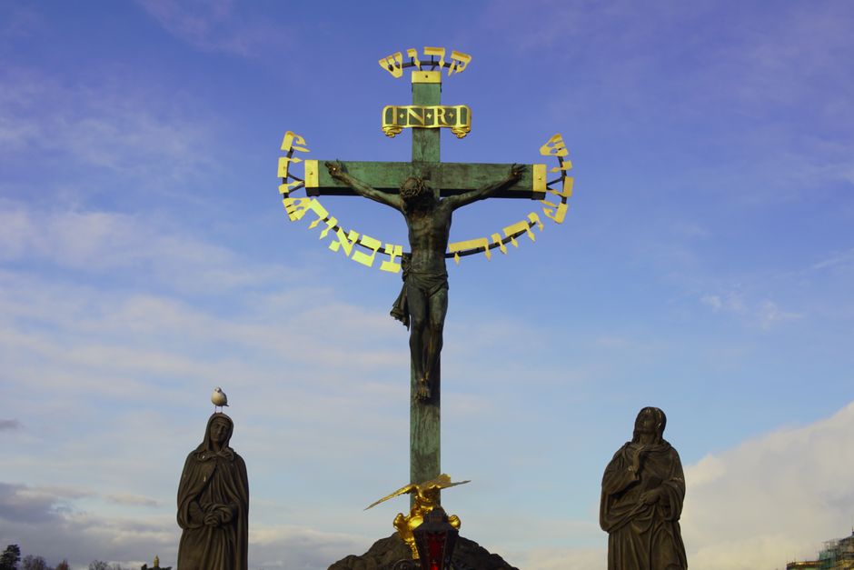 Estatua de crucifixión con inscripción en hebreo y dos figuras bajo un cielo vívido, ubicada al aire libre.