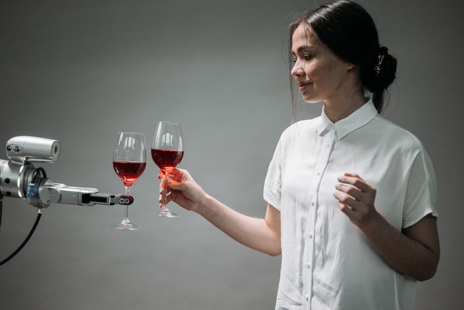 Una mujer y un brazo robótico chocan copas de vino tinto, simbolizando la armonía tecnológica.