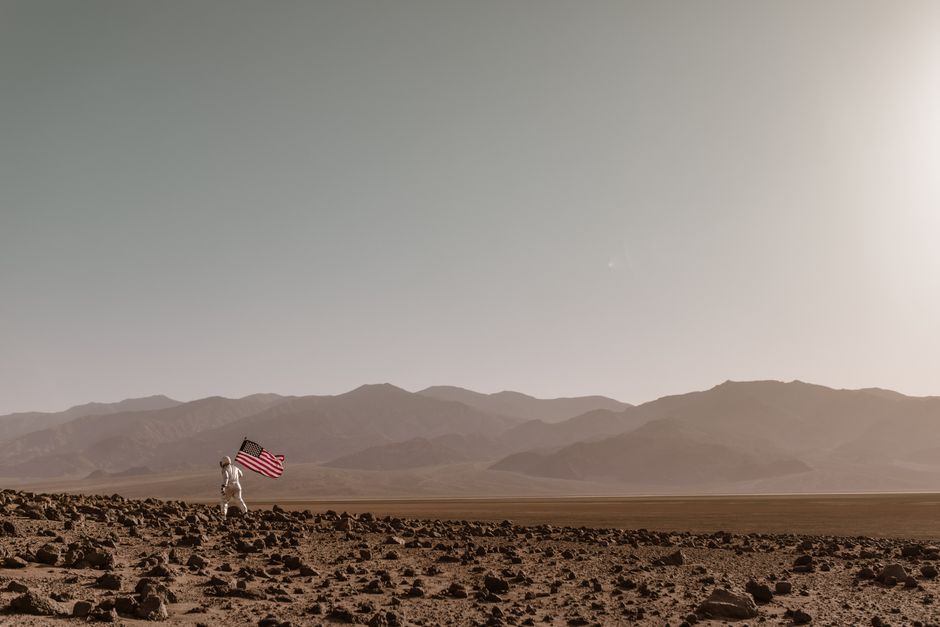 Astronauta en traje espacial caminando sobre un paisaje rocoso desolado sosteniendo una bandera americana.