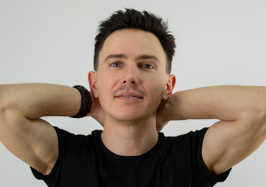 Retrato de un hombre seguro de sí mismo con cabello corto y negro, posando con las manos detrás de la cabeza contra un fondo neutro.