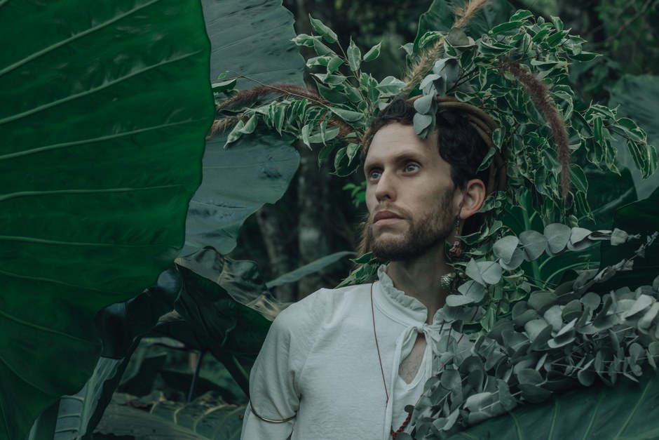 Hombre rodeado de frondoso follaje verde, adornado con elementos naturales, creando un aspecto etéreo.