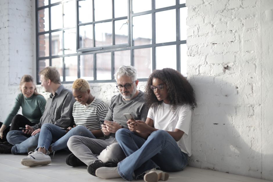 Un grupo diverso de adultos sentados en interiores junto a grandes ventanas, interactuando con smartphones.