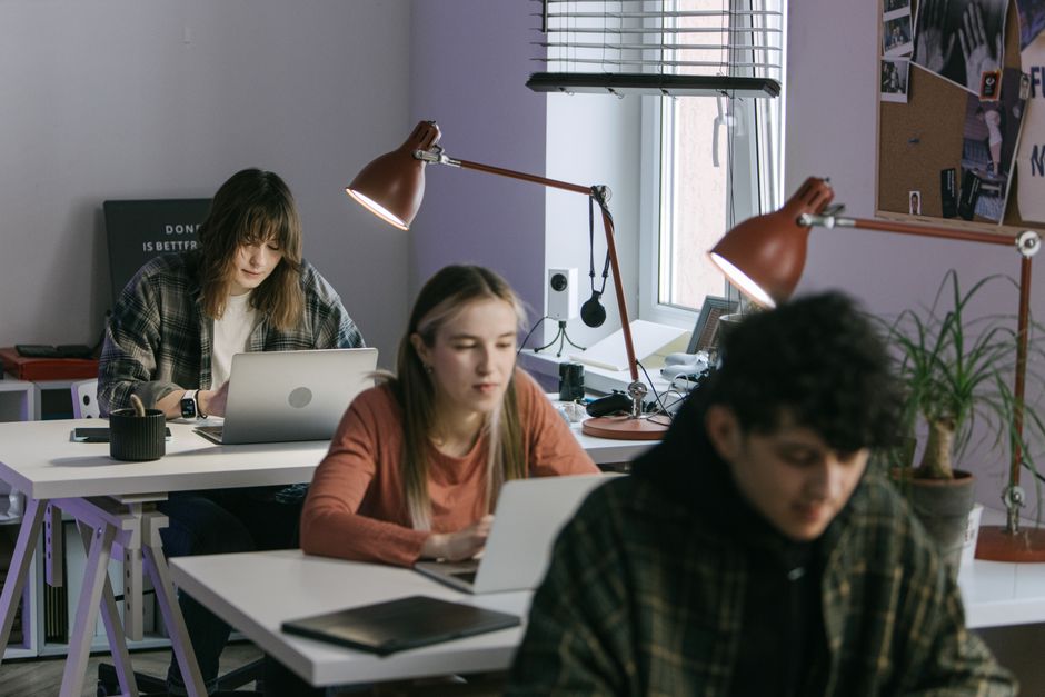 Tres jóvenes profesionales trabajando en escritorios en un entorno de oficina moderna con laptops y lámparas de escritorio.
