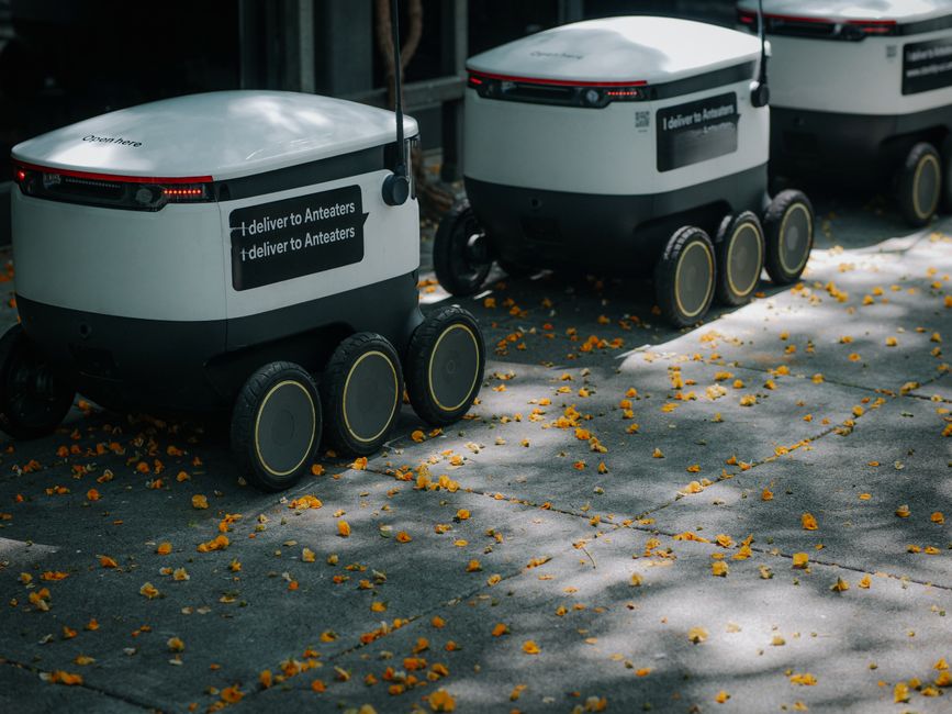 Robots de entrega futuristas navegando por una acera llena de hojas, mostrando innovación en la logística urbana moderna.