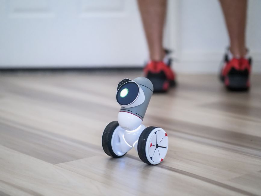 Un primer plano de un robot moderno con ruedas sobre un suelo de madera con piernas al fondo.