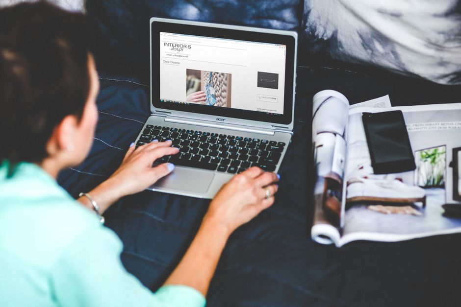 Una mujer en la cama navegando y blogueando con una laptop y una revista cerca.