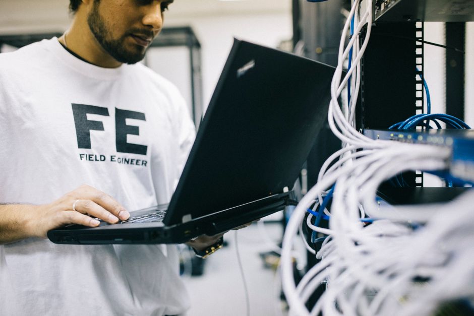 Un ingeniero de campo se concentra en el laptop mientras maneja cables de red en una sala de servidores.
