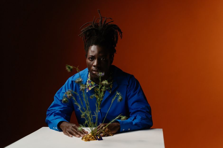 Un retrato conceptual de estudio de un hombre con estilo, con trenzas afro, vistiendo una camisa de mangas largas azules y sentado frente a un fondo naranja.