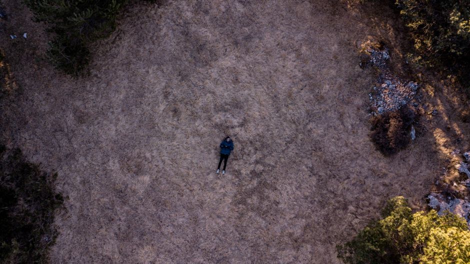 Una figura solitaria se encuentra acostada en un vasto campo abierto rodeado de naturaleza, capturada desde arriba.