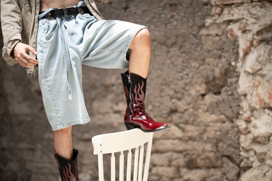 Una persona con estilo, de pie sobre una silla, luce prendas de mezclilla y botas únicas contra una pared texturizada.