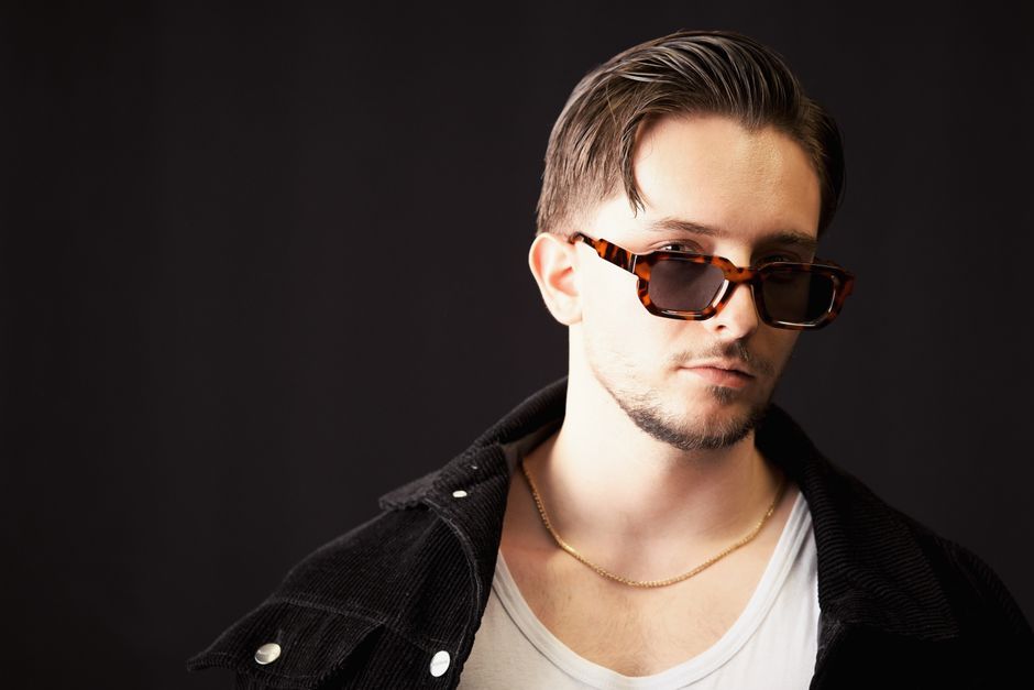 Retrato de un joven con gafas de sol contra un fondo oscuro, transmitiendo una vibra de moda y confianza.