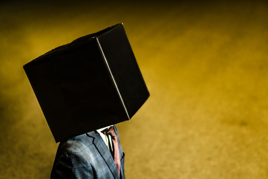 Una persona con chaqueta de traje está al aire libre con una caja negra en la cabeza contra un fondo amarillo.