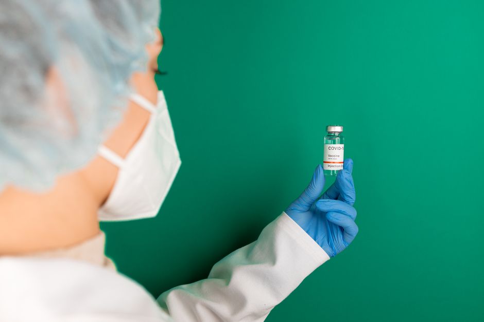 Un trabajador de la salud con guantes y mascarilla sostiene un vial de vacuna COVID-19 frente a un fondo verde.