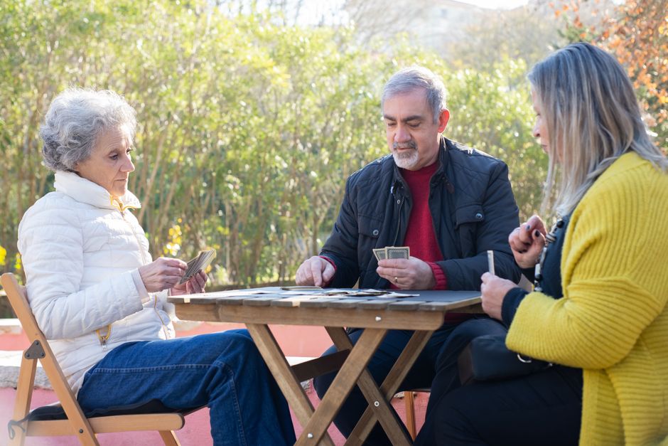 Tres personas mayores juegan a las cartas en una mesa de madera en un parque soleado, disfrutando de su tiempo de ocio al aire libre.