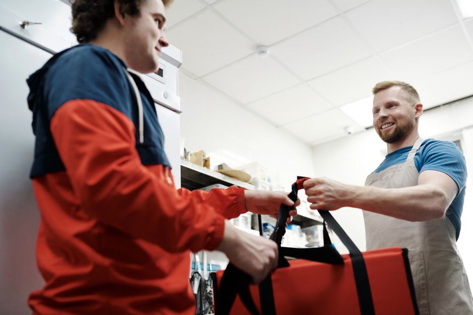 Trabajador sonriente le entrega una bolsa térmica a un repartidor en una cocina ocupada.