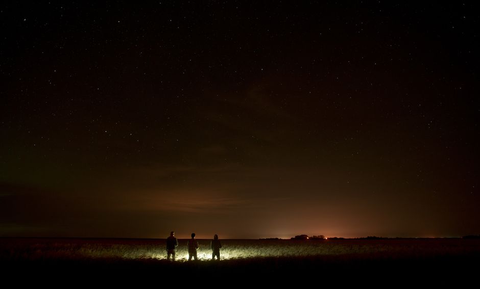 Tres personas bajo un cielo estrellado con luces de ciudad a lo lejos en el horizonte.