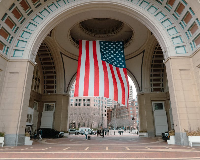 Una gran bandera estadounidense cuelga bajo un arco en Boston, capturando una escena urbana patriótica.