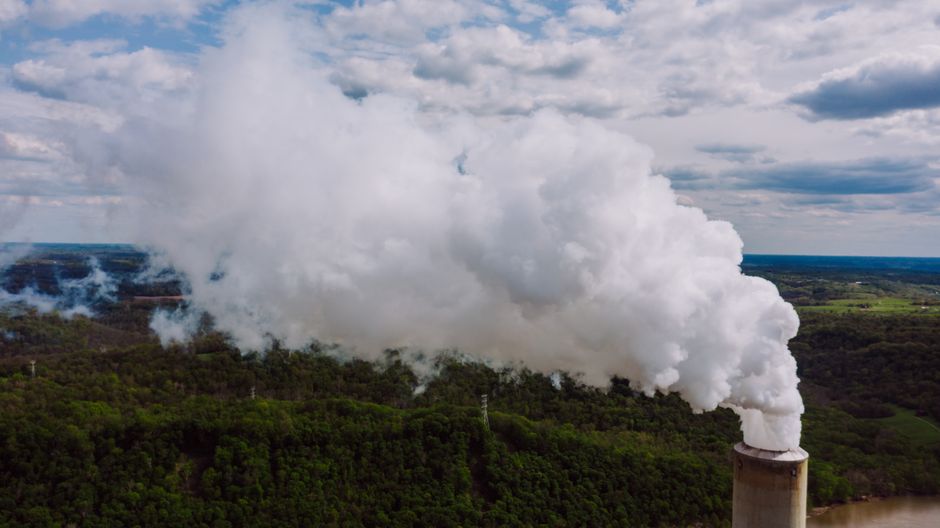 Toma de dron de una chimenea de planta energética emitiendo humo blanco espeso sobre un paisaje forestal.