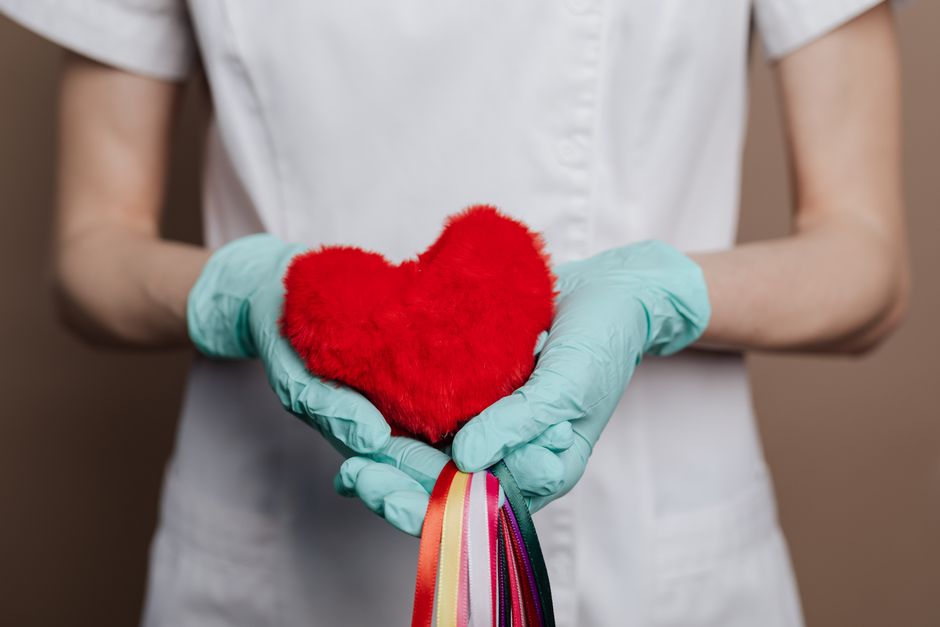 Una enfermera con guantes sostiene un corazón rojo con cuerdas de arcoíris, simbolizando amor y atención médica.