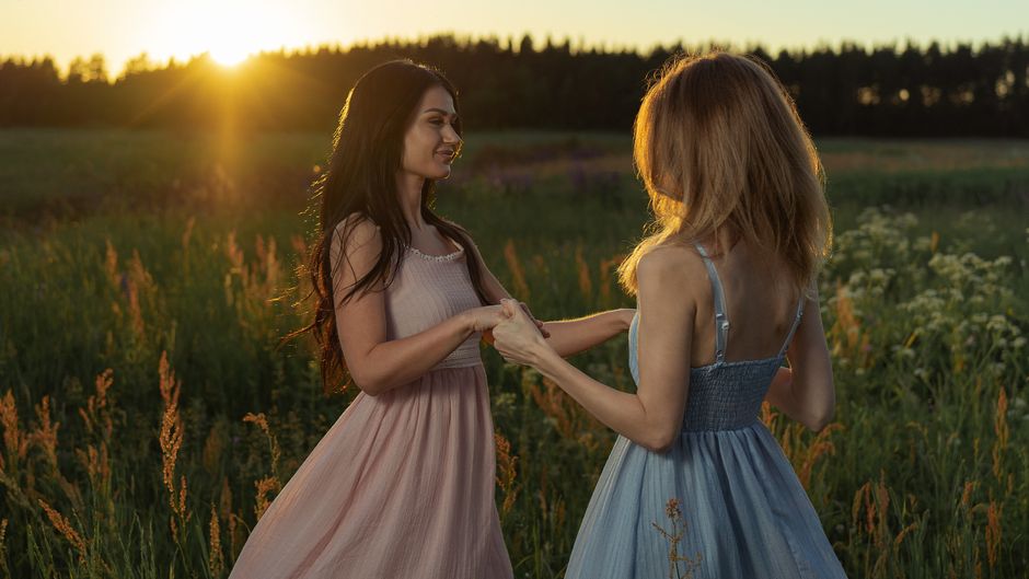 Dos mujeres en vestidos tomadas de la mano en un campo iluminado por el sol al atardecer.