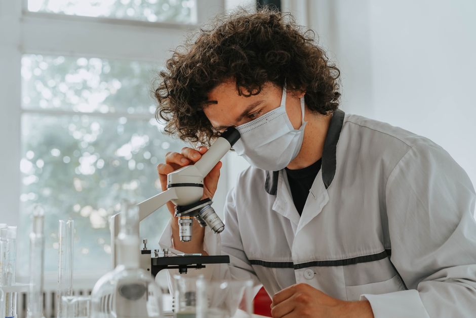 Un científico con mascarilla examina muestras bajo un microscopio en un entorno de laboratorio.