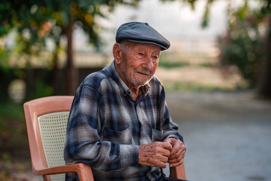Hombre adulto mayor sentado al aire libre, vistiendo una gorra y una camisa a cuadros, exudando tranquilidad.